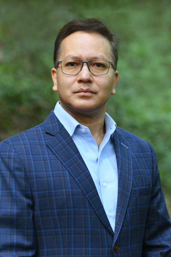Man in glasses wearing a blue checked blazer and light blue shirt outdoors.
